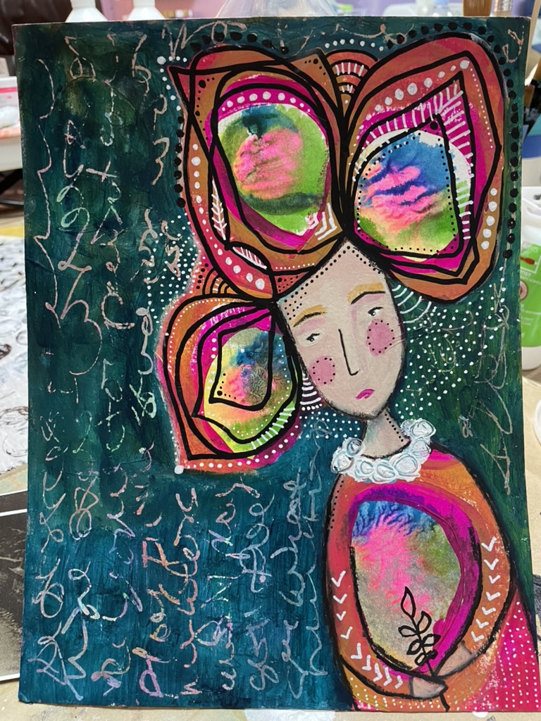 A folk-art portrait on a deep teal ground: a young woman with white skin and pink cheeks, holding a small painted bouquet, wearing a tall pink-orange headpiece patterned with green and pink shapes, surrounded by white script writing.