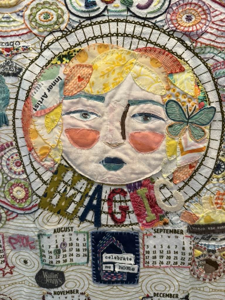 A textile portrait stitched from many small fabric scraps: a woman’s face with a halo of yellow and patterned petals, the word “MAGIC” spelled in mixed letters across the chest, with calendar pages and a small embroidered house at the bottom.
