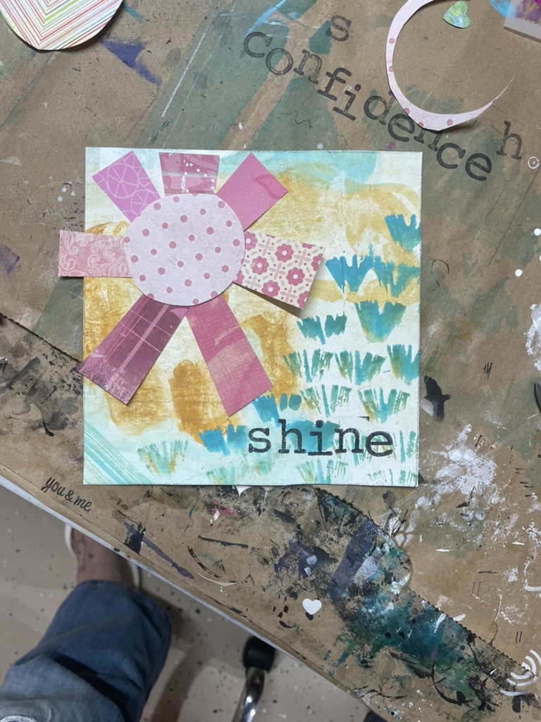 A small collage on a soft yellow-and-teal printed ground: pink ribbons radiate from a polka-dotted paper sun, with the word “shine” stamped in black at the lower right.